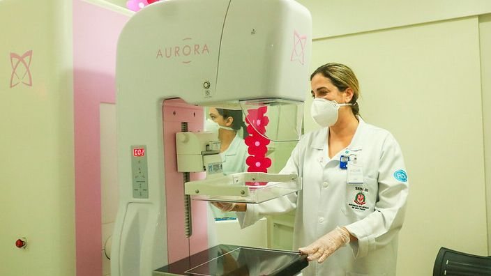 Mamografia a cada dois anos é indicada após os 50 anos, a depender do caso #pratodosverem Foto colorida com duas mulheres de jaleco e máscara brancos. Uma está ao fundo da sala branca com uma parede rosa. A outra mulher está na frente de um mamógrafo - Divulgação PMB