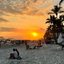 Imagem mostra um fim de tarde com o sol brilhando intensamente e colorindo o céu de tons dourados e laranjas na faixa de areia de uma praia urbana com coqueiros e prédios ao fundo