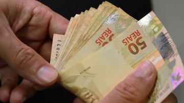 Mão segura cédulas de dinheiro e representa o pagamento do seguro-desemprego
