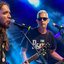 Uma banda em apresentação ao vivo, com guitarra, baixo, saxofone e vocais, sob iluminação em tons de azul