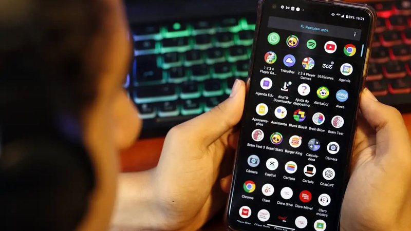 smartphone com vários aplicativos abertos na tela enquanto observa o aparelho; ao fundo, um teclado iluminado aparece desfocado em ambiente interno