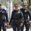 A imagem mostra três guardas civis municipais em uniforme escuro e coletes à prova de balas parados ao lado de uma viatura azul e amarela, em uma rua movimentada e ensolarada