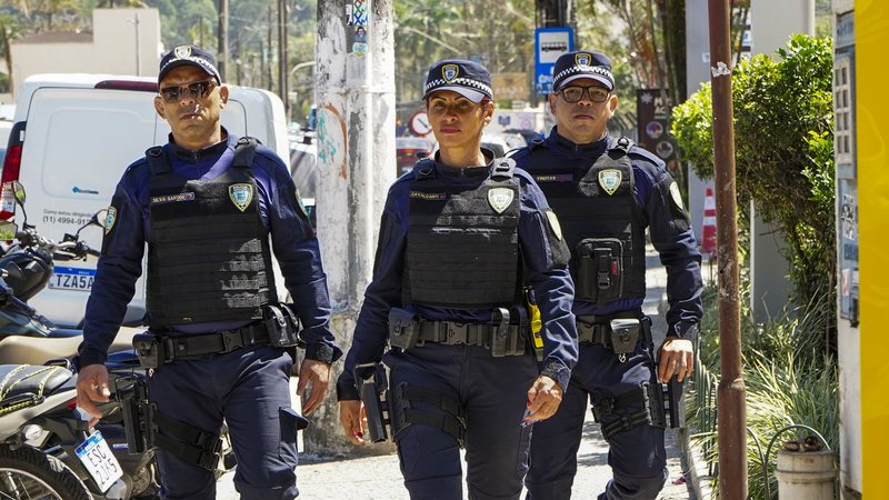 A imagem mostra três guardas civis municipais em uniforme escuro e coletes à prova de balas parados ao lado de uma viatura azul e amarela, em uma rua movimentada e ensolarada