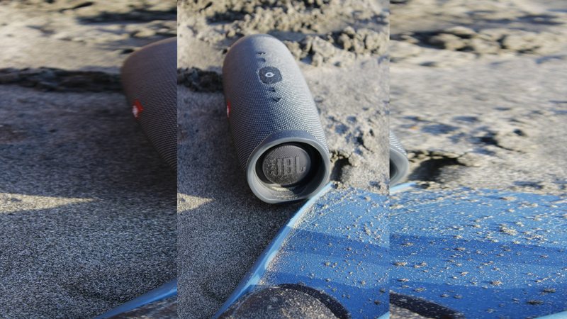 Caixa de som portátil cilíndrica apoiada na areia da praia ao lado de uma prancha azul