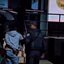 A imagem mostra uma pessoa algemada sendo conduzida por um agente de segurança fardado para o interior de uma viatura policial caracterizada da Polícia Militar