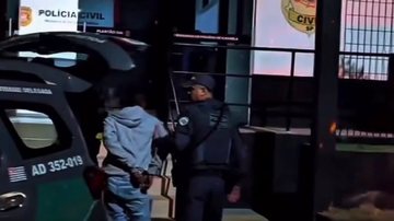 A imagem mostra uma pessoa algemada sendo conduzida por um agente de segurança fardado para o interior de uma viatura policial caracterizada da Polícia Militar