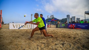 A imagem mostra uma partida de beach tennis (ténis de praia) a decorrer numa faixa de areia à beira-mar, com atletas concentrados no jogo e edifícios urbanos ao fundo