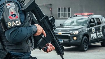 Imagem mostra um policial militar do BAEP (Batalhão de Ações Especiais de Polícia) da Polícia Militar do Estado de São Paulo (PMESP) com um fuzil e uma viatura ao fundo
