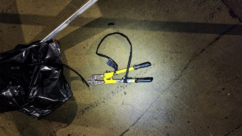 Imagem mostra um alicate crimpador hidráulico manual (ou prensa-terminais) de corpo amarelo com cabos emborrachados pretos, apoiado no chão de concreto ao lado de um saco plásticos preto e um pedaço de cabo elétrico ou fio