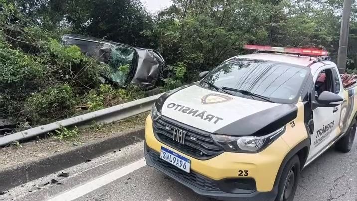 Imagem mostra uma viatura do trânsito de São Sebastião (SP) em uma rodovia onde um veículo menor aparece acidentado e fora da pista, encostado no talude de vegetação