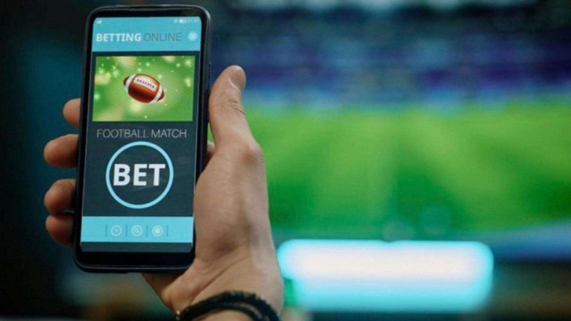 Imagem mostra uma mão segurando um smartphone com uma tela genérica de aplicativo de apostas esportivas ("bet") com um campo de futebol e um botão "BET"