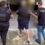 Imagem mostra uma pessoa detida sendo escoltada por dois agentes, um deles vestindo um colete tático com a identificação do DENARC (Departamento Estadual de Investigações sobre Entorpecentes) e outro com uma camisa preta da Polícia Civil