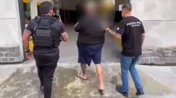 Imagem mostra uma pessoa detida sendo escoltada por dois agentes, um deles vestindo um colete tático com a identificação do DENARC (Departamento Estadual de Investigações sobre Entorpecentes) e outro com uma camisa preta da Polícia Civil
