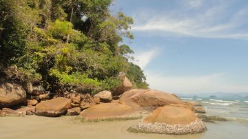 Praia das Bicas, com areia clara, grandes rochas à beira-mar, vegetaçãoe o oceano ao fundo