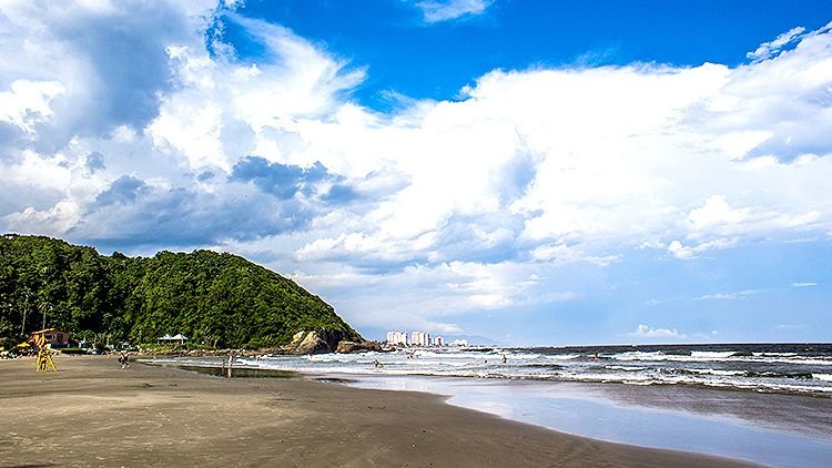 Praia de Itanhaém com faixa de areia extensa, mar agitado, vegetação e morro ao fundo