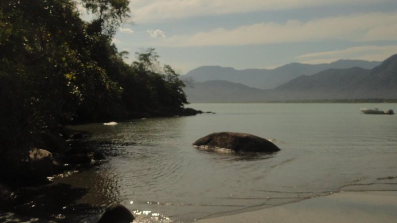Praia Estaleiro do Padre, com águas tranquilas, vegetação, pedras e montanhas ao fundo