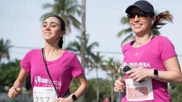 Imagem mostra duas participantes correr juntas numa prova de corrida de rua ao ar livre, vestindo camisolas cor-de-rosa com o logotipo "Run Riviera".