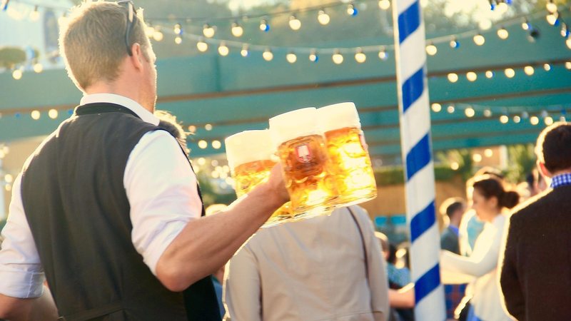 Imagem mostra um garçom transportando várias canecas grandes de cerveja num ambiente festivo ao ar livre, típico de um festival como a Oktoberfest