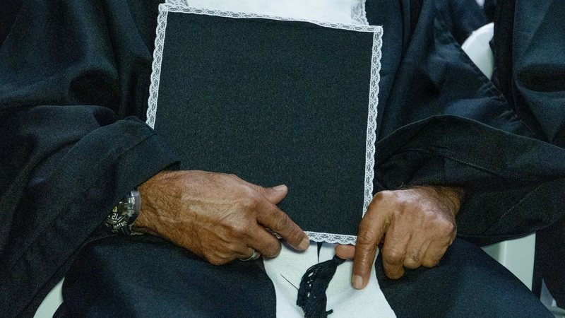 Homem sentado e vestido com uma beca segurando uma capela