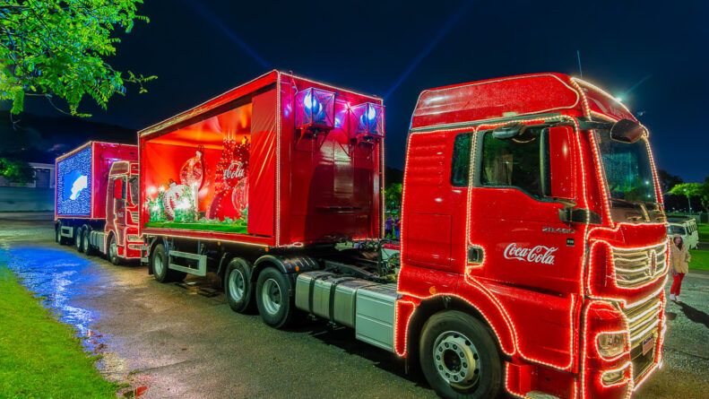 A imagem mostra os tradicionais caminhões vermelhos iluminados da Caravana de Natal da Coca-Cola
