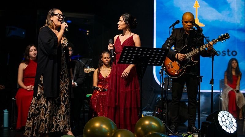 A imagem mostra músicos e cantoras se apresentando ao vivo no palco durante um evento musical