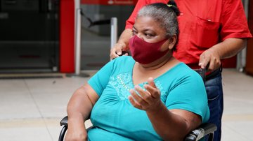 Campanha de tampinhas e lacres beneficia mulher com cadeira de rodas em Guarujá Gueomar Leda sentada na nova cadeira de rodas e atrás dela o esposo dela com as mãos na cadeira - Divulgação/Prefeitura de Guarujá