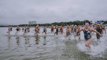 Imagem de competição realizada em anos anteriores Bertioga tem retomada do esporte com volta do Performance Run Mulheres de maiôs, óculos e toucas correndo e entrando no mar para uma competição de travessia aquática - Arquivo/Prefeitura de Bertioga