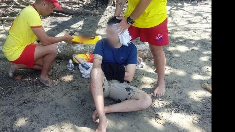 Turista sofre queda em pedras de praia em Ilhabela  Turista ferido no chão com dois salva vidas ao lado - Reprodução