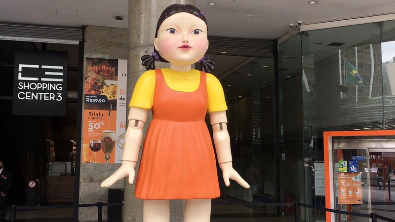 boneca vestida de laranja e amarelo montada na entrada do shopping Round 6: boneca sinistra rouba cena na avenida mais badalada de São Paulo Boneca gigante vestida de laranja e amarelo na entrada do shopping - Reprodução/Twitter