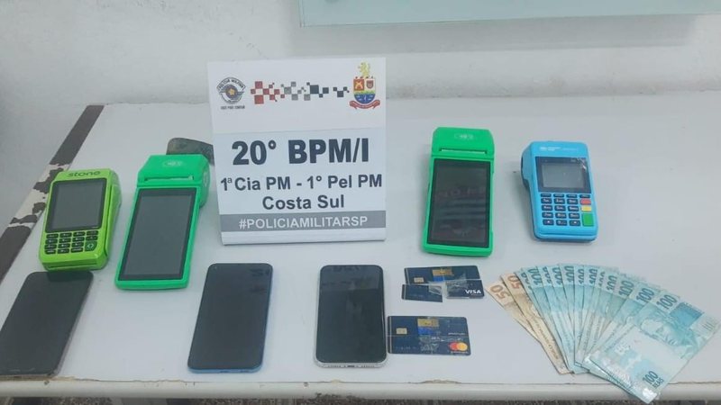 Máquina de cartão, dinheiro e celulares apreendidos PM prende criminosos que aplicavam ‘golpe do cartão’ em São Sebastião (SP) dinheiro, celurares e maquinas de cartão sobre a mesa - Foto: Vigésimo BPMI