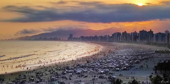 Litoral de SP deve registrar altos índices de calor, diz Climatempo Tempo no litoral praia lotada de banhistas, com pôr do sol de cores alaranjadas - Hebe Dorado/Facebook