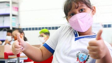 Estudante de Itanhaém com máscara. Item voltou a ser obrigatório em escolas municipais da cidade Uso de máscaras volta a ser obrigatório em escolas municipais de Itanhaém Menina com máscara dando joia - Imagem: Divulgação / Prefeitura de Itanhaém