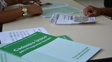Serão distribuídas 150 senhas por ordem de chegada na Vila do Bem Centro Sábado (24) tem mutirão do CadÚnico em Bertioga Papelada do CadÚnico em mesa, onde pessoa é atendida ao fundo - Prefeitura de Bertioga