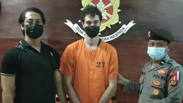 Jovem está 'morando' desde o dia 28 de junho no aeroporto de Bali; parentes relatam precariedade Brasileiro detido na Indonésia Dois agentes da Indonésia ao lado esquerdo e direito e jovem de laranja detido no meio - Divulgação