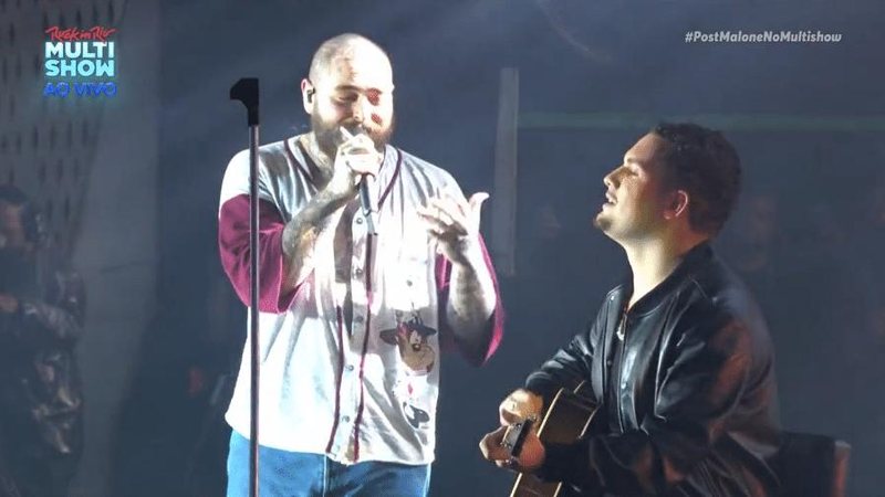 Theo Kant é morador de Niterói (RJ) e conseguiu ficar na grade do show para mostrar seu cartaz e ter a oportunidade de subir ao palco Post Malone Post Malone, rapper careca e com um microfone nas mãos com o fã tocando violão no palco - Reprodução