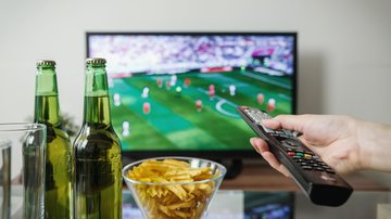 A Globo vai transmitir 56 das 64 partidas da Copa Copa do Mundo: veja onde assistir aos jogos do Mundial do Catar Pessoa assistindo TV com jogo de futebol, segurando controle remoto e cerveja e petiscos na mesa em frente - Pexels