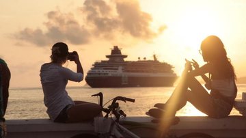Primeiro navio chega na quarta-feira (2) Santos: temporada de cruzeiros prevê 561 mil turistas Pessoas fotografando navio de passageiros chegando à cidade durante pôr do sol - Prefeitura de Santos