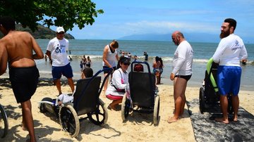 Pessoas com mobilidade reduzida visitam o mar Caraguatatuba retoma programa Praia Acessível Duas pessoas com deficiência estão sentadas nas cadeiras anfíbias que estão na areia. Uma mulher conversa com uma delas. Outras pessoas estão em pé e outras na água - Imagem: Divulgação / Cláudio Gomes / Prefeitura de Caraguatatuba