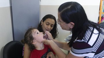 As crianças devem estar acompanhadas dos pais Bertioga leva campanha de vacinação para escolas municipais Criança no colo da mãe com a boca aberta tomando vacina - Divulgação/Prefeitura de Bertioga