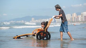 Na temporada, os atendimentos serão desenvolvidos de terça-feira até domingo, das 10 às 16 horas Programa Praia Acessível Homem levando uma criança em um equipamento para o mar - Divulgaçãço
