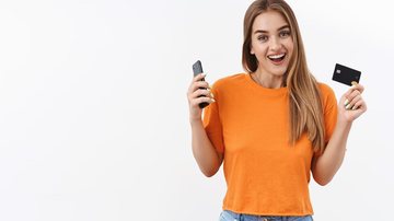 Mulher loira vestida com uma camiseta laranja e calça diz segurando um celular e um cartão de credito - Freepik