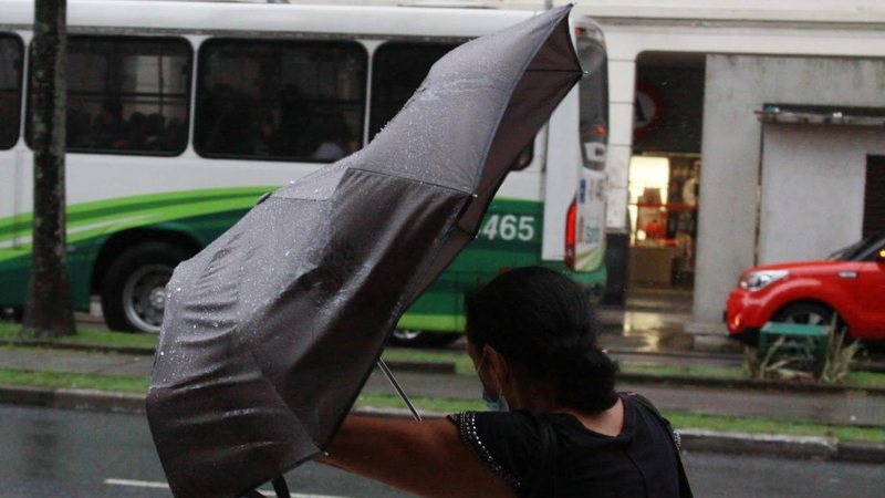 Defesa Civil pede cautela no locais perto de prédios em construção, onde a força do vento pode fazer com que materiais sejam arremessados Previsão do tempo indica fim de semana de fortes ventos e chuva em Santos Mulher com guarda-chuva envergando pelo vent - Prefeitura de Santos